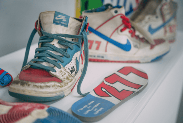 Worn, vintage sneakers with red, blue, and white accents are displayed on a shelf. One shoe has a teal lace, and next to it lies a colorful insole featuring a "77" design. Nearby, another sneaker with a bold blue Nike swoosh is visible.